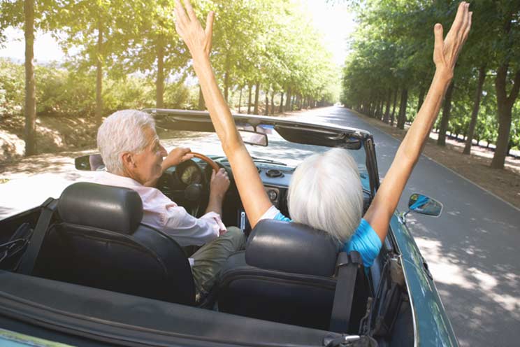 an excited elderly couple driving a convertible with the woman in the passenger seat extending her arms up