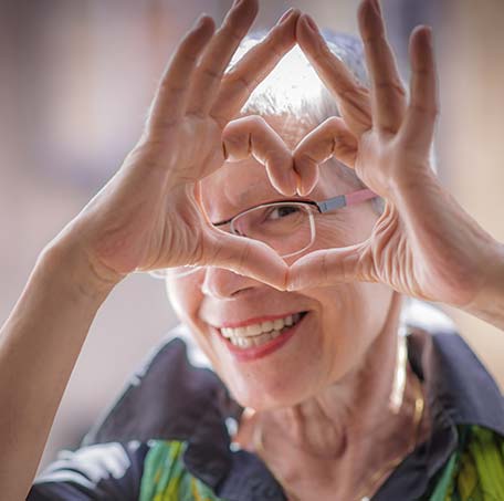 a smiling elderly woman peeking through a heart shape she is making with her hands